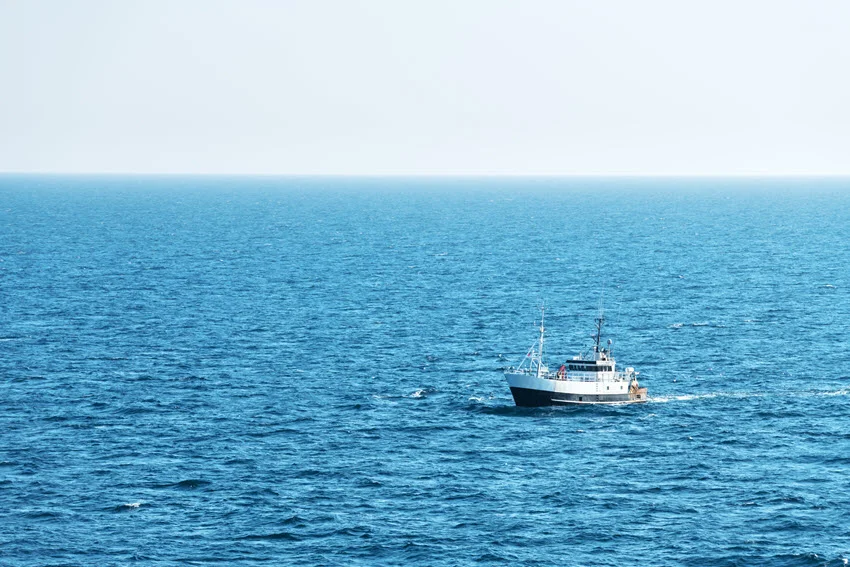 Distance view of a fishing boat drifting across open blue water beneath a clear sky.