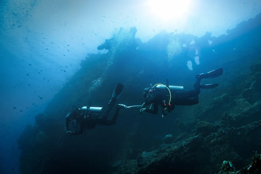 An underwater shot from below the surface, featuring two divers exploring an old shipwreck amidst coral reefs, with light visible from above.