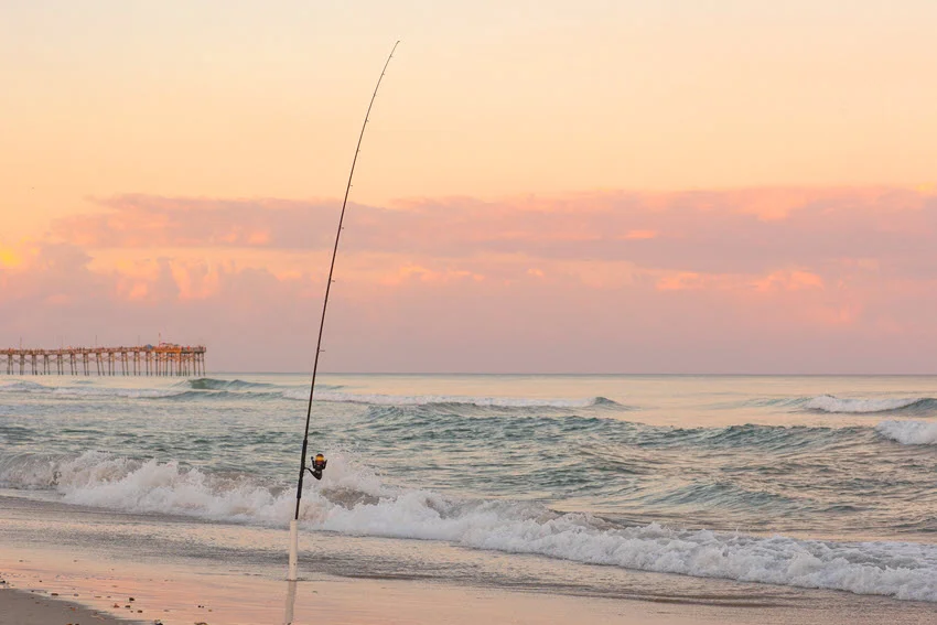 Fishing pole in the surf on the beach with a pier in the background.