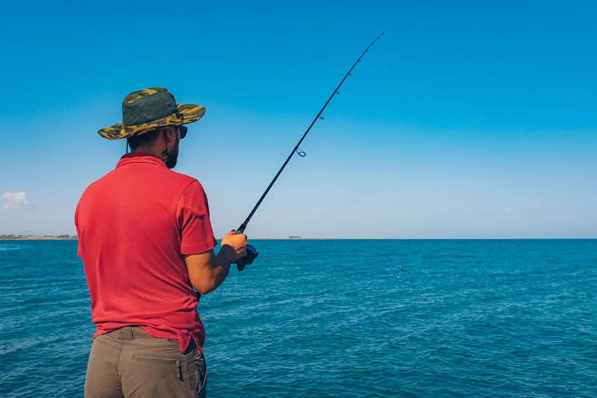 Back shot of an angler standing on the shore, casting a line into the ocean under clear blue skies on a bright summer day.
