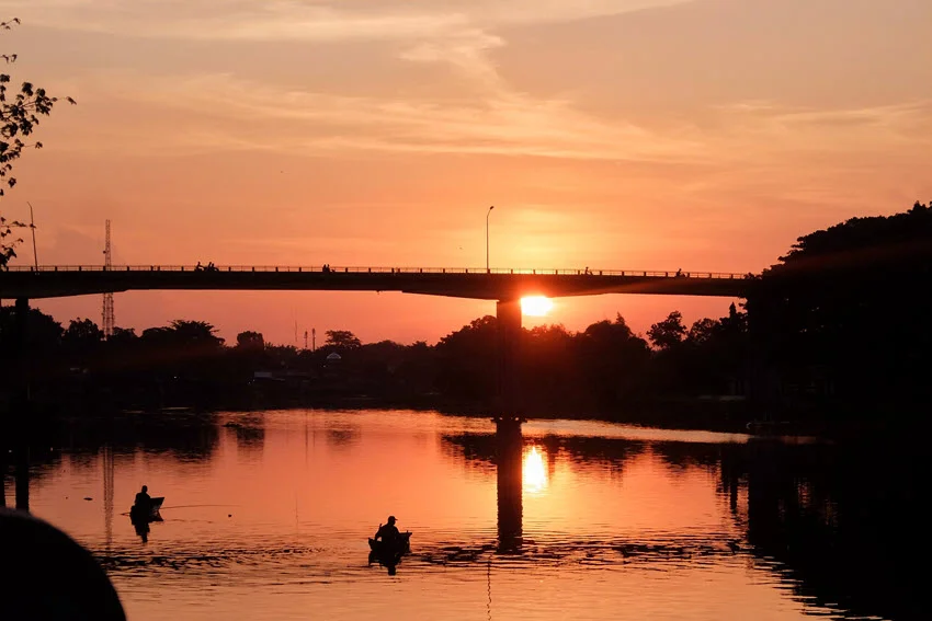 Two smaller fishing boats drift in the river below the bridge during a magnificent sunset.