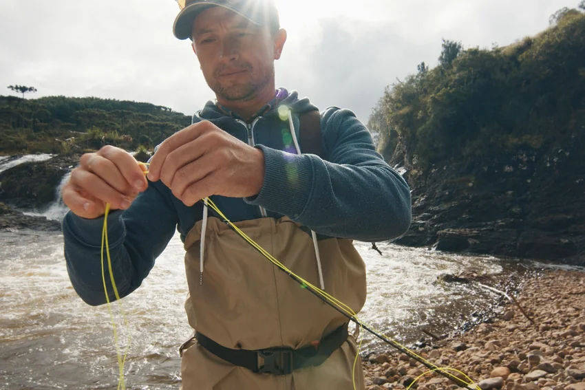 Beside a rocky river, an angler in waders handles a bright fly line with both hands while a rod rests across the body.