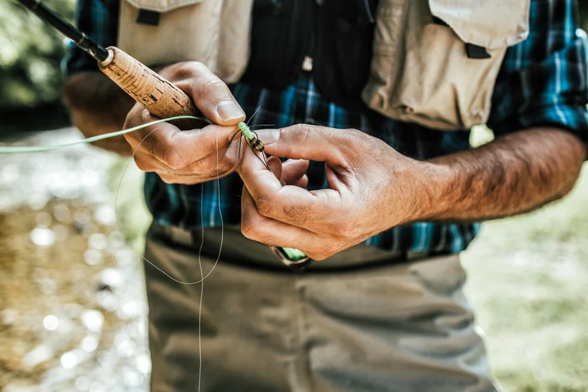 An angler holds an artificial fly and thin line beside a cork rod handle, with a fishing vest and checkered shirt visible.