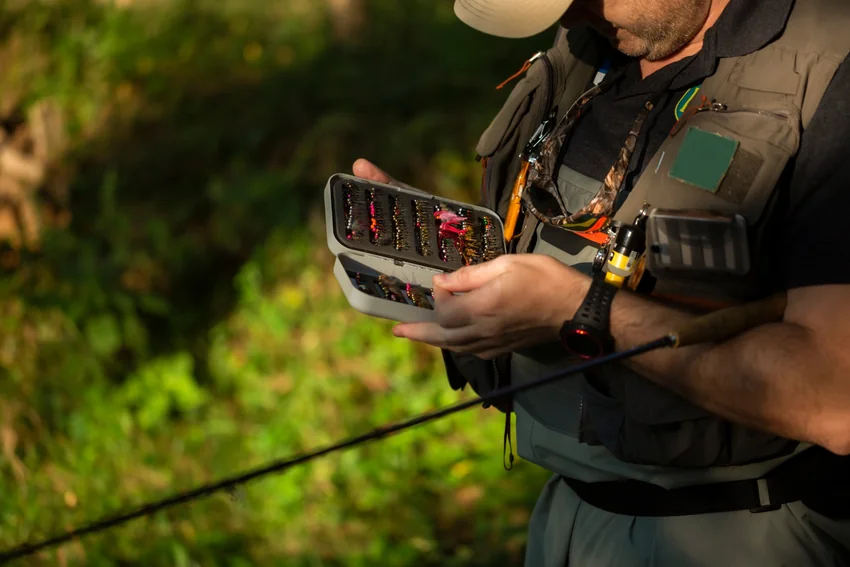 An angler wearing a fishing vest examines an open fly box beside a rod, with rows of artificial flies visible inside.