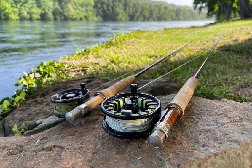 Two fly rods with cork handles and reels lie on a rocky riverbank, with flowing water and trees in the background.