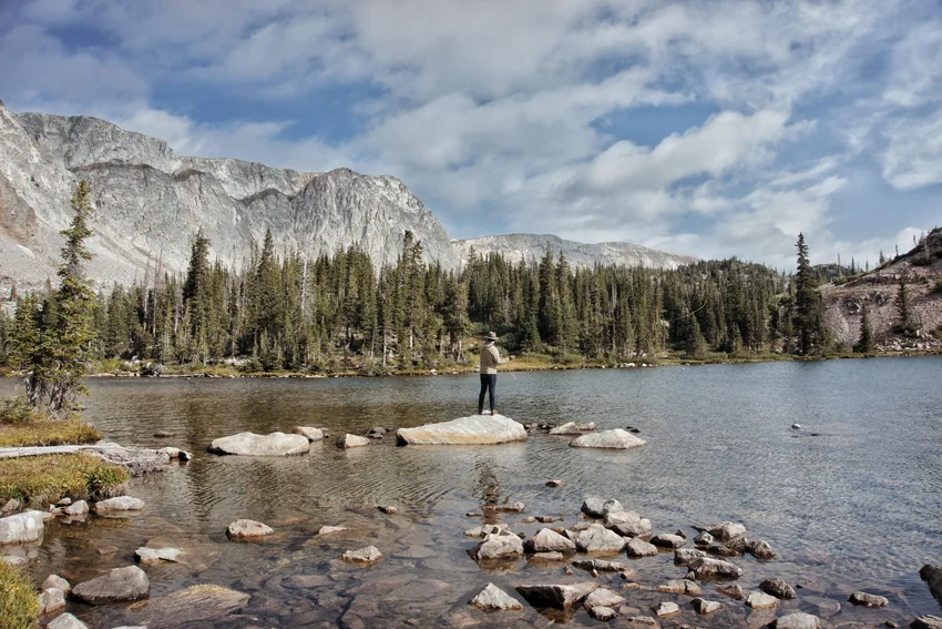 An angler stands on rocks in a mountain river, surrounded by a rocky shoreline, evergreen trees, distant peaks, and a cloudy sky.