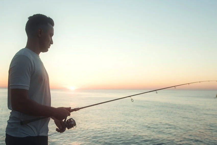 An angler with a fishing rod stands on the shore and enjoying a beautiful sunset.