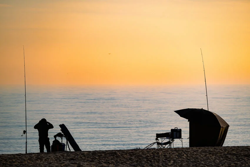 A man is fishing on the beach, equipped with a tent, fishing gear, and surf rods stuck in the sand.