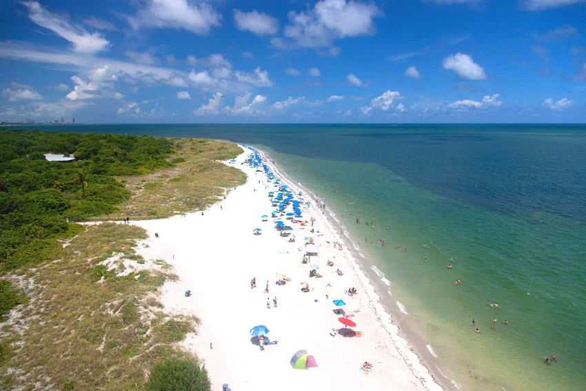 Aerial view of the long white sandy beach at Bill Buggs Park with many beachgoers during a beautiful sunny day.
