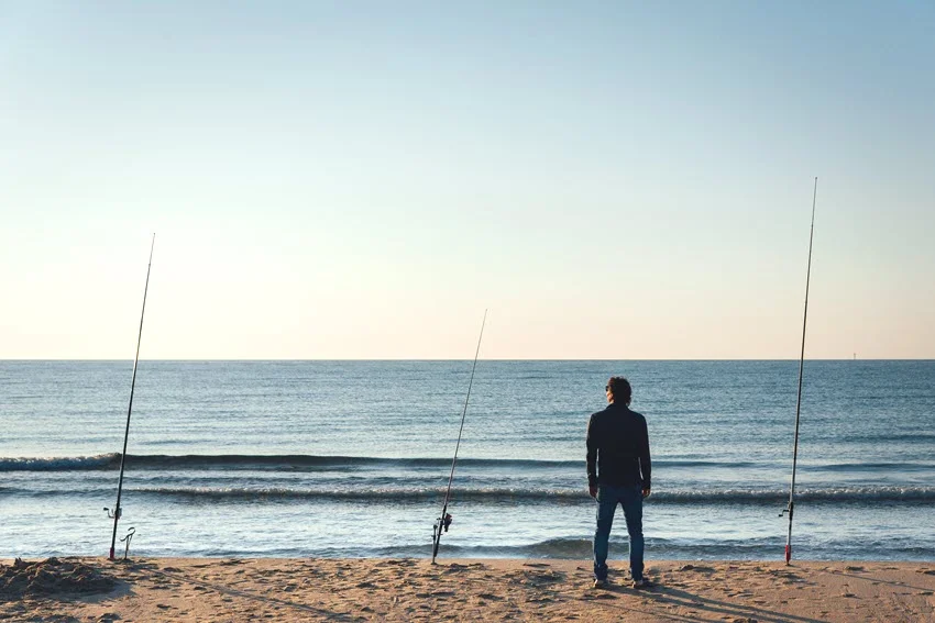 An angler stands beside several surf-fishing rods planted in the sand while watching the water from the shoreline.