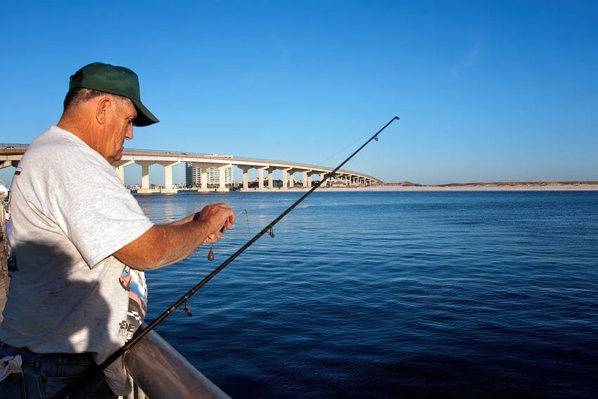 An angler prepares bait beside the water at Florida Point, with a fishing rod and bridge visible behind him.