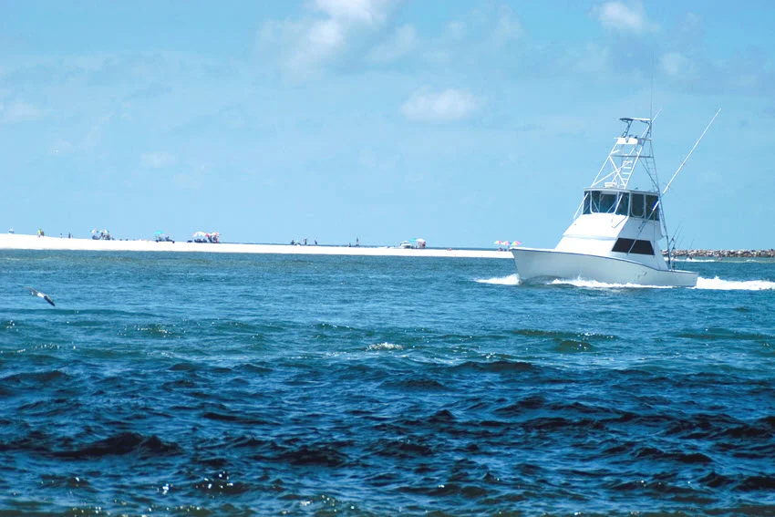 An offshore fishing charter boat crosses Gulf water near Orange Beach, with a low sandy shoreline visible beyond the rolling waves.