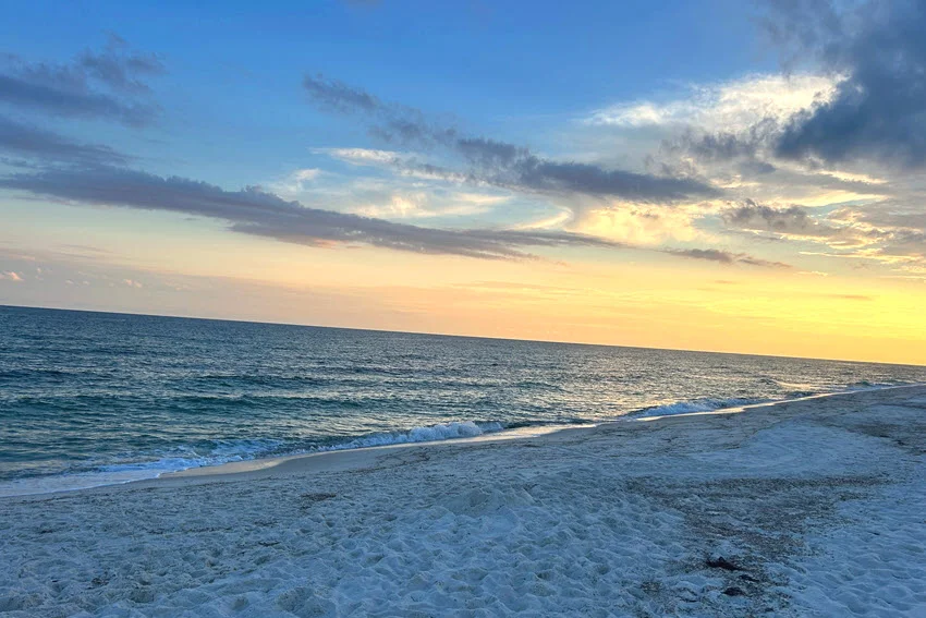 Sunset colors spread across the Orange Beach shoreline as gentle waves reach the sand beneath a calm evening sky.