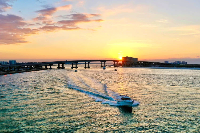 A fishing charter boat travels through Perdido Pass beside the Orange Beach shoreline under a clear sky and calm coastal water.