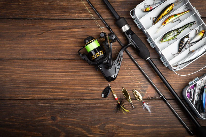 Top down view of a spinning rod, reel, hooks, lures, and tackle boxes are arranged on a wooden surface for an overview of basic fishing gear.
