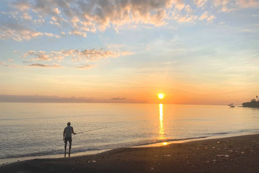 Back view of an angler casting from the beach into calm inshore waters during sunset.