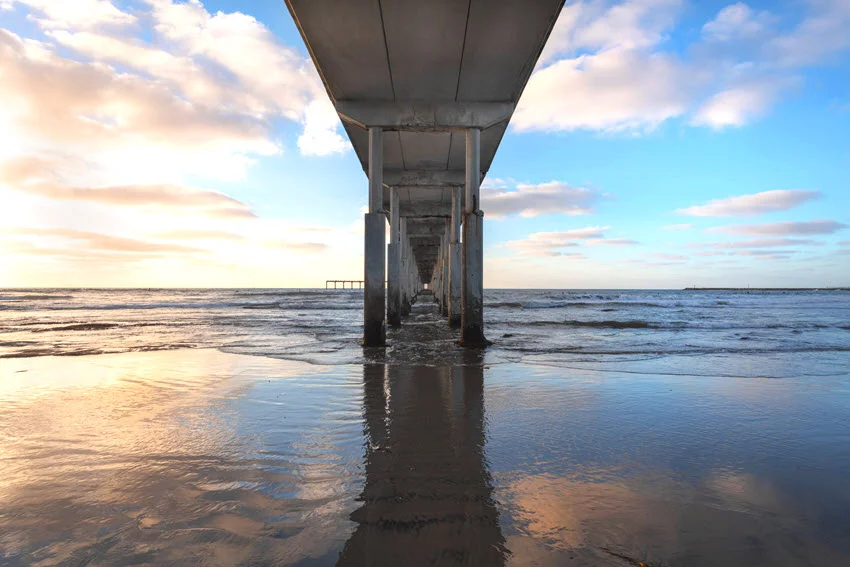 View of a fishing pier from the shore below at sunset with warm colors reflecting across the sky and water.