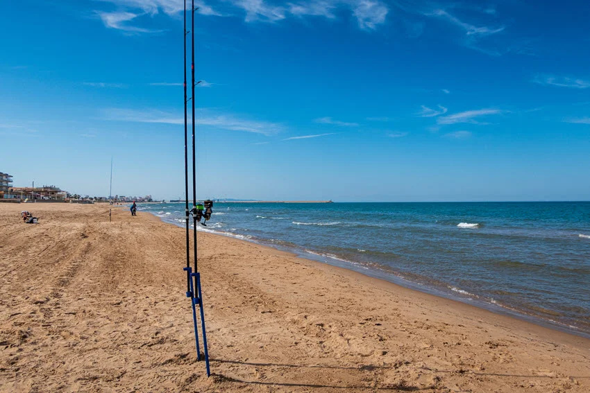 View of two surf fishing rods on the beach with a few anglers in the distance casting into the surf.