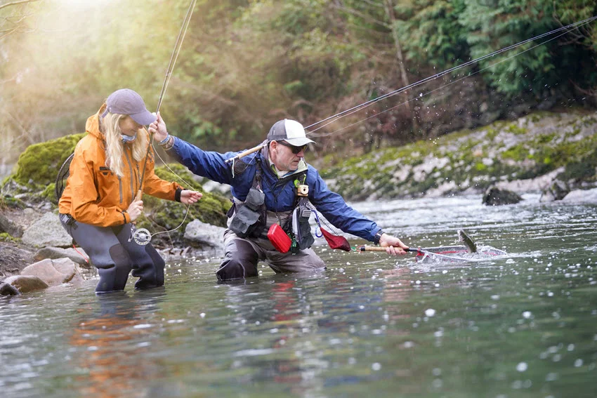 Guide reaching with a landing net as a woman fly fishes in a fast, rocky stream surrounded by dense forest.