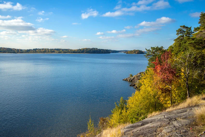 Wide view of a calm freshwater lake bordered by rocky shoreline and autumn-colored trees beneath a bright blue sky.