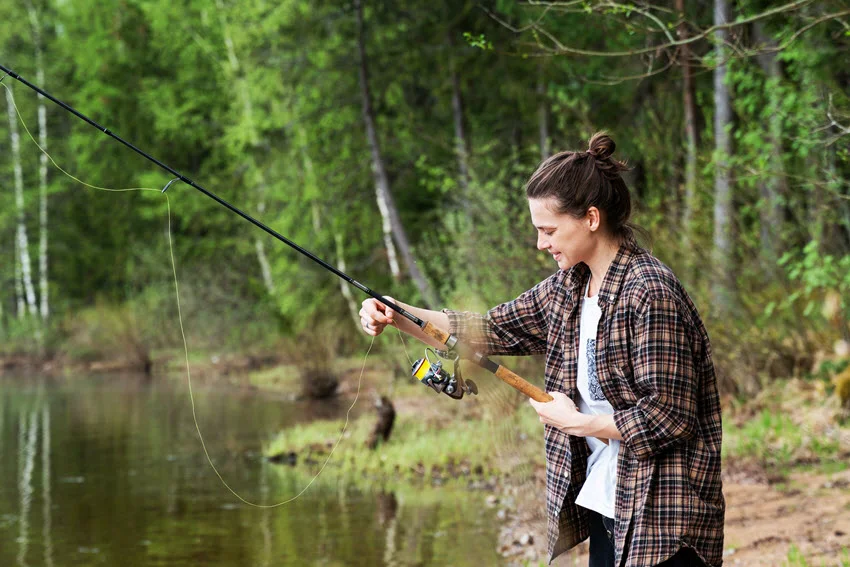 Woman preparing a spinning rod beside a wooded pond, demonstrating a simple shore-fishing setup for freshwater game fish.