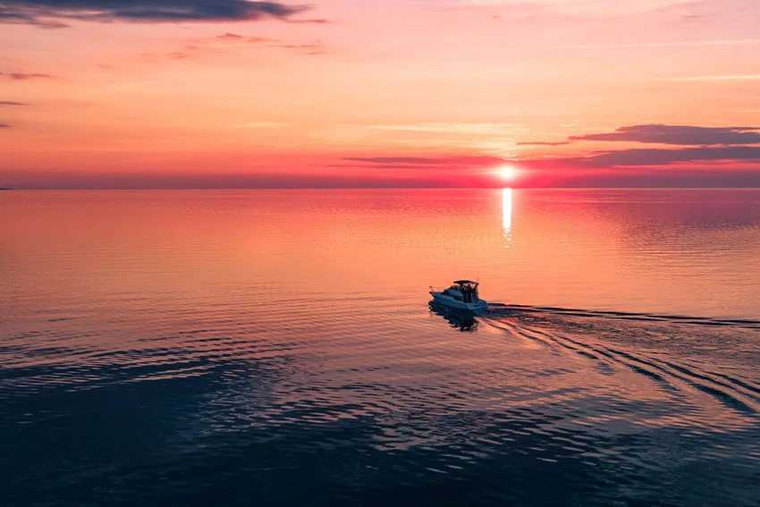 Boat crossing Chaumont Bay, New York, at sunset, with calm Lake Ontario water reflecting warm pink and orange light.
