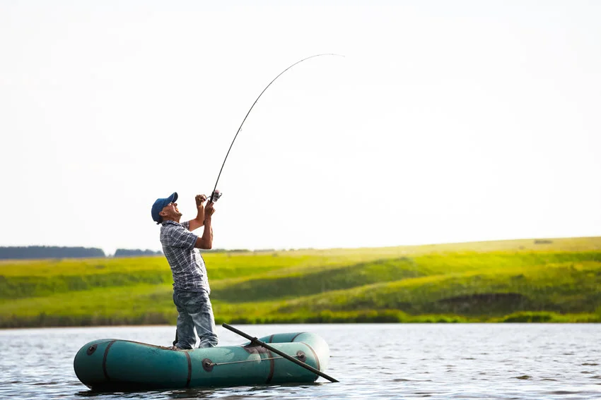 Angler drifting for Perch from a small inflatable boat on open lake water, with green fields stretching across the distant shoreline.