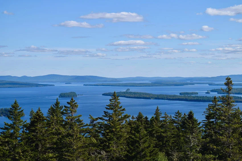 Wide view of Lake Cobbosseecontee, Maine, surrounded by evergreen forest, distant hills, and small islands beneath a clear blue sky.