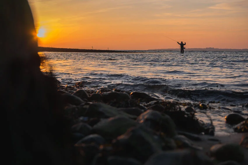 Silhouette of an angler swinging a fly fishing rod in the ocean during sunset.