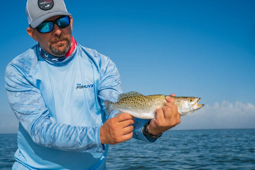 A close-up shot of an angler holding a Speckled Trout with both hands on a boat during a sunny day, wearing a scarf around the neck styled like the American flag.