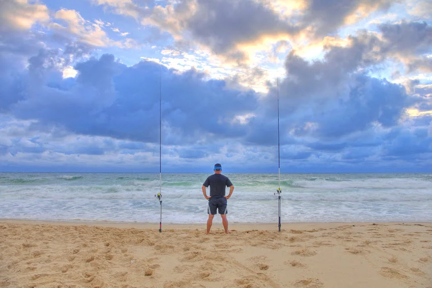 A lone surf angler stands on a sandy beach between two fishing rods in a dusk waits for  fish to bait.