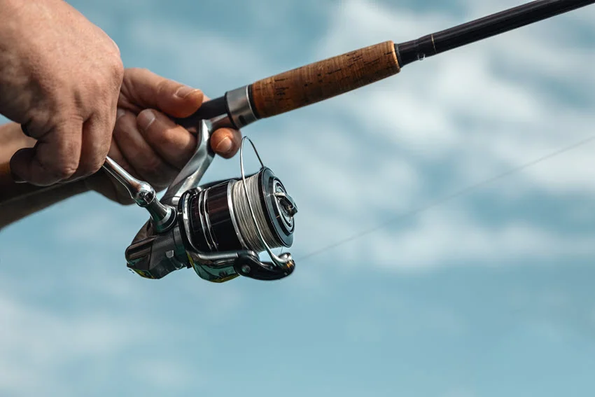 Focused shot of freshwater fishing in summer, showing a spinning reel mounted on a spinning rod held by a fisherman, with a blurred background of greenery and water.