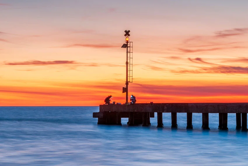 A pier at sunset with two fishermen wearing headlamps as they prepare their gear.