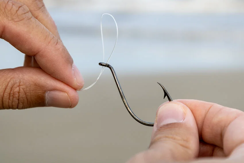 An angler's hands threading the line through the hook, with a blurred beach shore in the background.