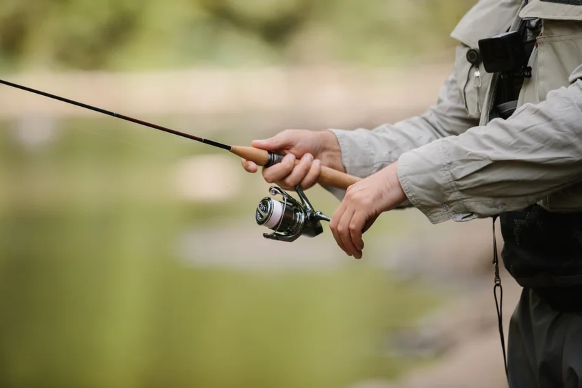 Angler wearing a gray jacket and fishing vest holds a spinning rod and reel beside the water, with both hands on the tackle.