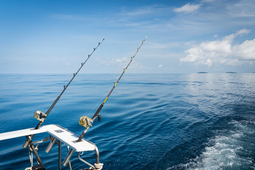 Two fishing rods and reels sit in holders at a boat's stern, with lines extending over the sea beside its wake.