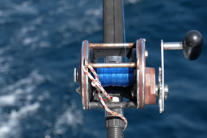 Conventional fishing reel filled with blue line sits on a rod above the sea, with metal fittings and the handle visible.