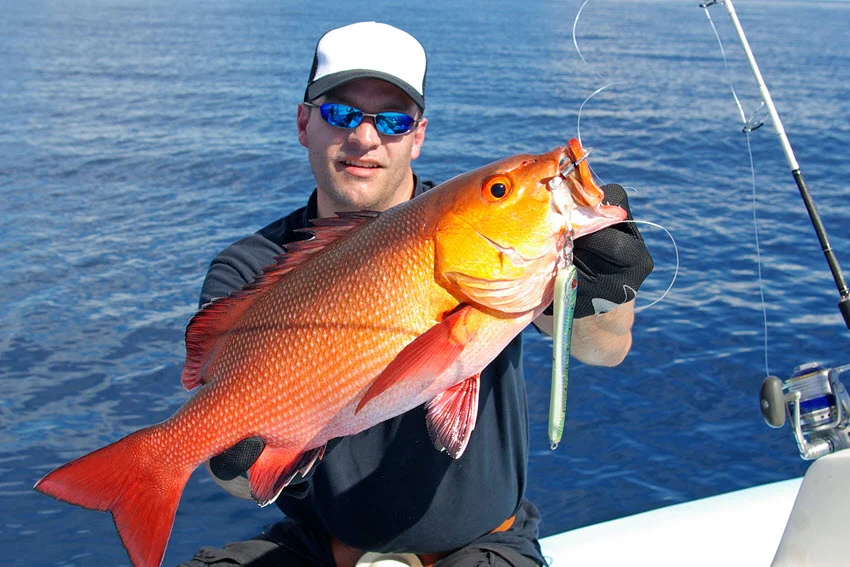 An angler holds a large Red Snapper aboard a boat, with a metal jig hanging beside its open mouth and blue water behind.