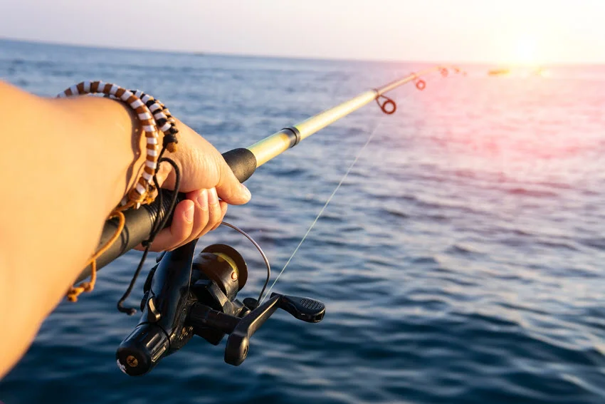 An angler grips a spinning rod over the sea, with the reel in the foreground and warm sunlight spreading across the water.