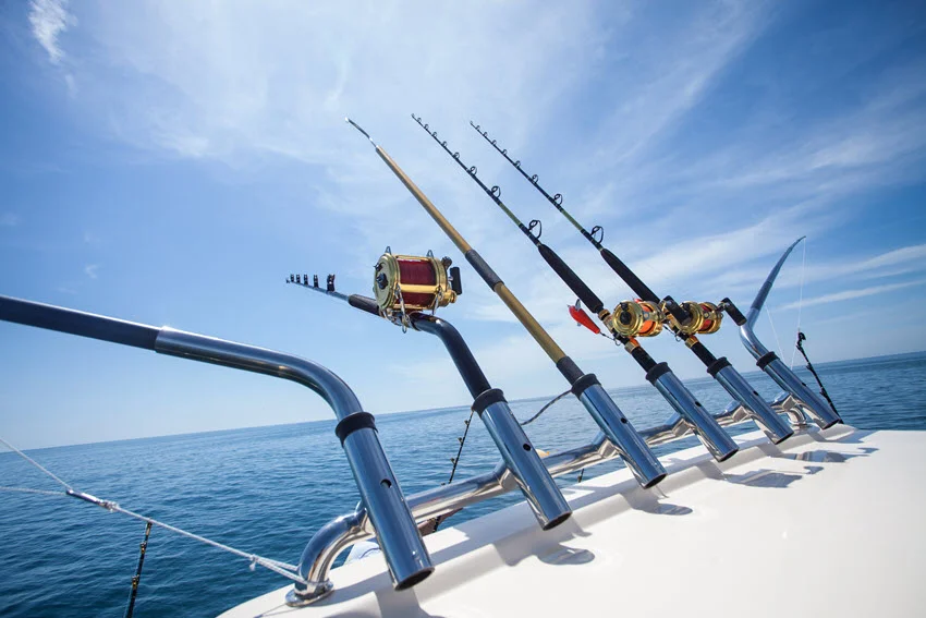 Big-game fishing rods with gold conventional reels stand in holders along a boat's stern, with calm ocean water beneath wispy clouds.