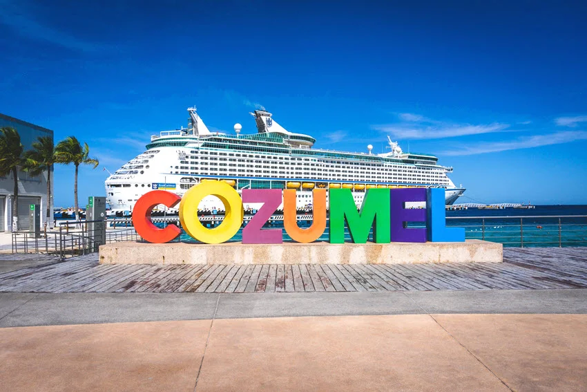 Colorful Cozumel letters stand on the waterfront in Mexico, with a large cruise ship docked behind them beside blue Caribbean water.
