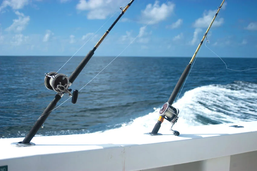 Two rods with conventional reels sit in holders along a boat's rail, with foamy wake trailing behind during an offshore fishing trip.