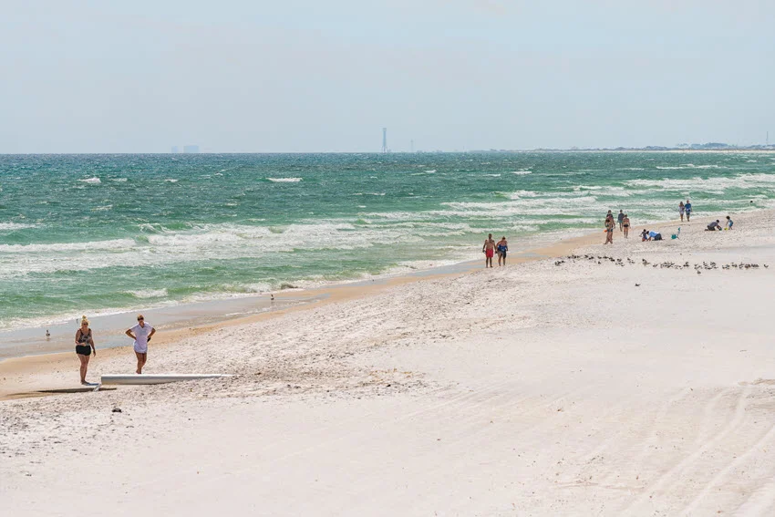 People walk along the white sand of Okaloosa Island near Fort Walton Beach, Florida, as waves roll ashore beside resting shorebirds.