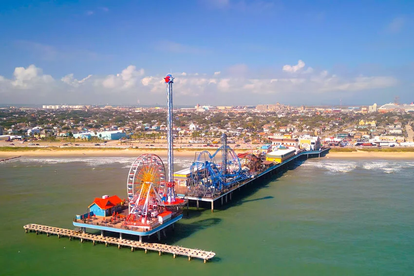 Galveston's Pleasure Pier extends over Gulf waters, with a Ferris wheel, roller coaster, and other rides backed by the Texas shoreline.