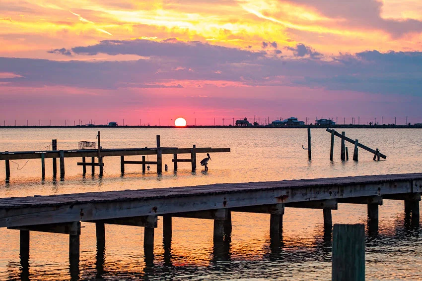The setting sun colors Caminada Pass at Grand Isle, Louisiana, pink and gold, with weathered docks and pilings silhouetted across the water.