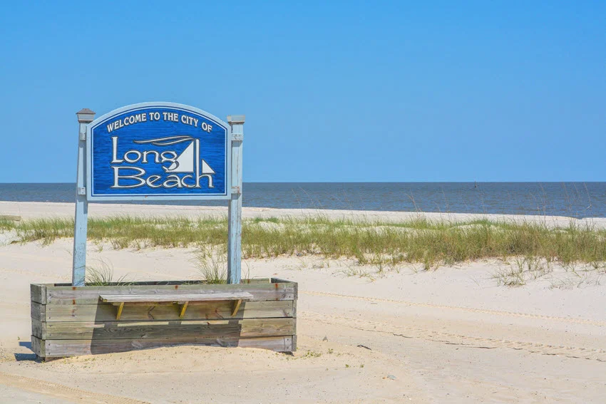 A blue welcome sign marks Long Beach, Mississippi, beside white sand and dune grass, with Mississippi Sound stretching across the background.