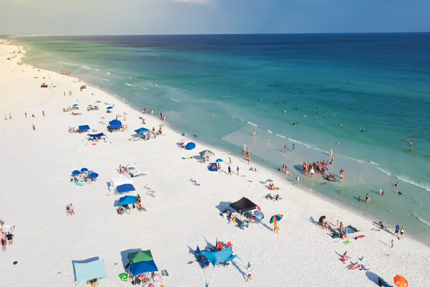 Beachgoers gather beneath colorful umbrellas on Miramar Beach near Destin, Florida, while swimmers enjoy the turquoise water along the white-sand shore.