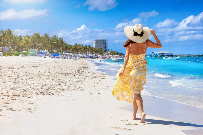 A woman in a yellow dress and sunhat walks along a Nassau beach in the Bahamas, beside turquoise water and palm-lined sand.
