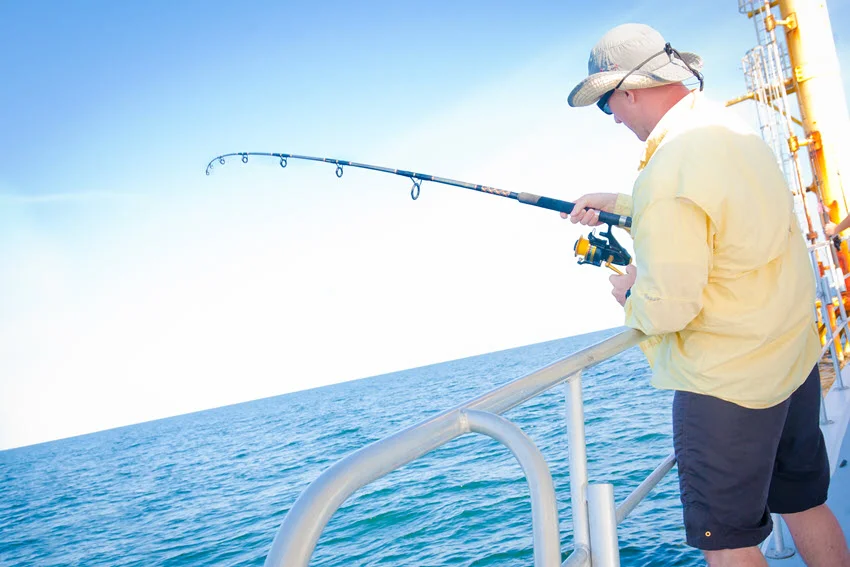 An angler in a yellow shirt reels from an offshore boat, holding a bent spinning rod over the rail above blue water.