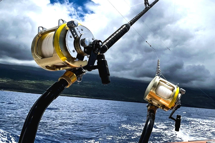 Two large gold conventional reels sit on mounted rods aboard a boat, with a forested coastline beneath heavy clouds across the water.
